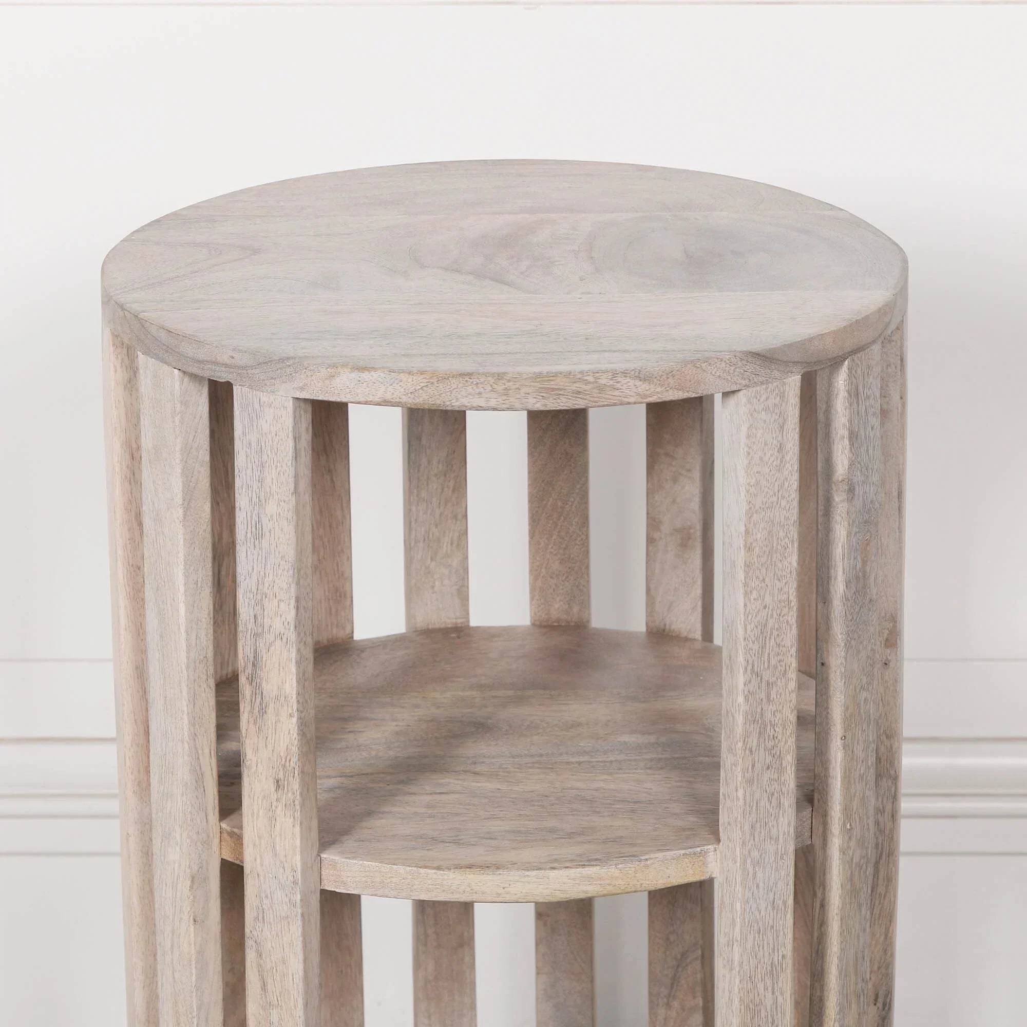 Light wood round side table with slatted base and two shelves against white wall