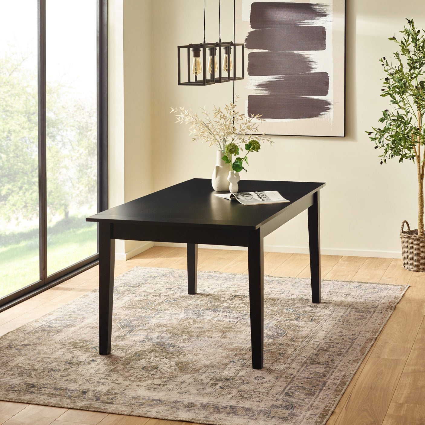 Black rectangular dining table with tapered legs, on patterned beige rug near large window and light wall art