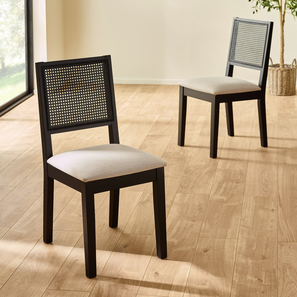 Two black wood dining chairs with woven cane backs and beige seats on light wood floor by large window.