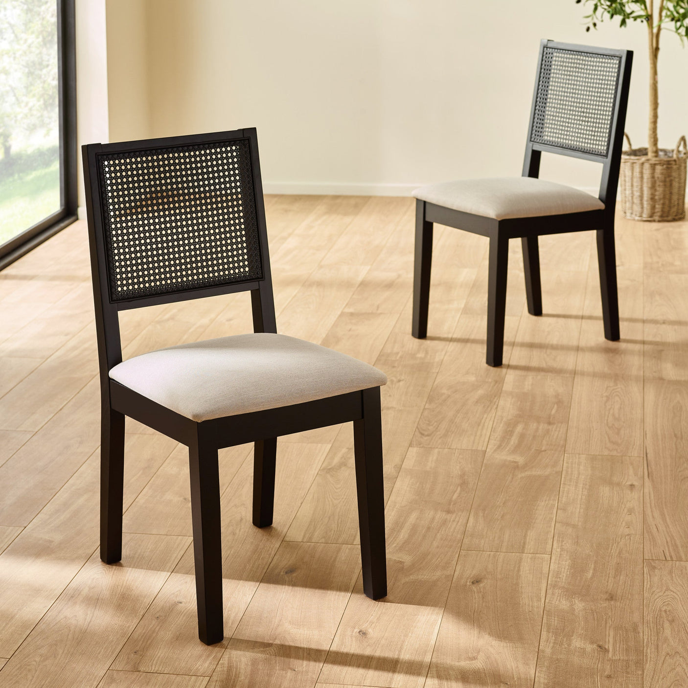 Two black wood dining chairs with woven cane backs and beige seats on light wood floor by large window.