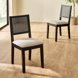 Two black wood dining chairs with woven cane backs and beige seats on light wood floor by large window.
