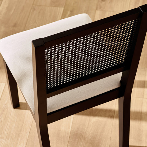 Rear angle of black dining chair showing woven cane backrest and light beige cushioned seat on wood floor