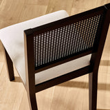 Rear angle of black dining chair showing woven cane backrest and light beige cushioned seat on wood floor