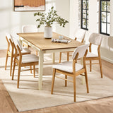 Rectangular dining table with natural wood top and cream legs with six white cushioned wooden chairs in a bright room.