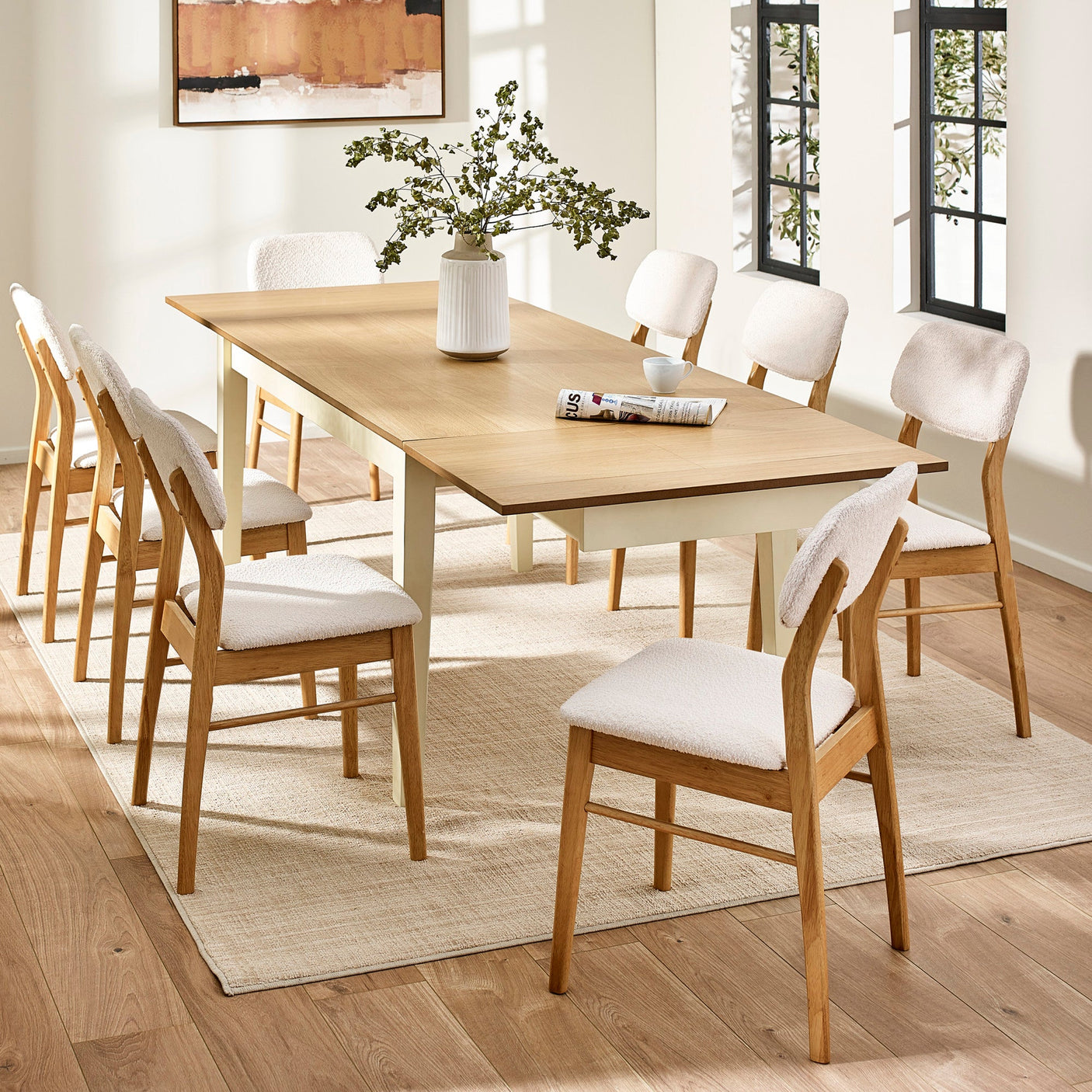 Extended rectangular dining table with cream legs and natural wood top and eight white cushioned wooden chairs in a bright