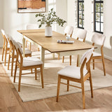 Extended rectangular dining table with cream legs and natural wood top and eight white cushioned wooden chairs in a bright