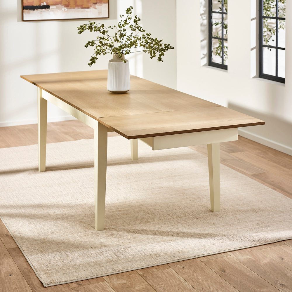 Extending rectangular dining table pulled out with light wood top and cream legs on beige rug by windows