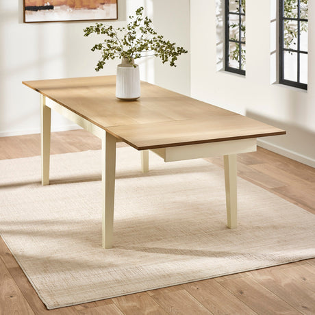 Extending rectangular dining table pulled out with light wood top and cream legs on beige rug by windows