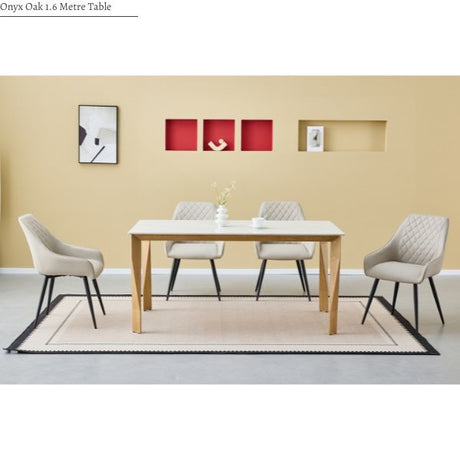 A dining room scene showing a rectangular Oak 1.6 metre table with a light wood finish and four upholstered dining chairs in a light beige color with black legs, arranged on a patterned rug against a beige wall.