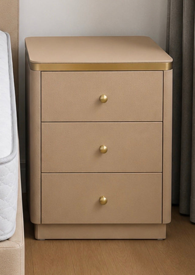 A three-drawer nightstand in a warm beige tone with a gold metal trim along the top edge and three round gold drawer knobs, shown in a bedroom setting next to a bed.
