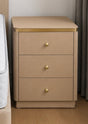 A three-drawer nightstand in a warm beige tone with a gold metal trim along the top edge and three round gold drawer knobs, shown in a bedroom setting next to a bed.