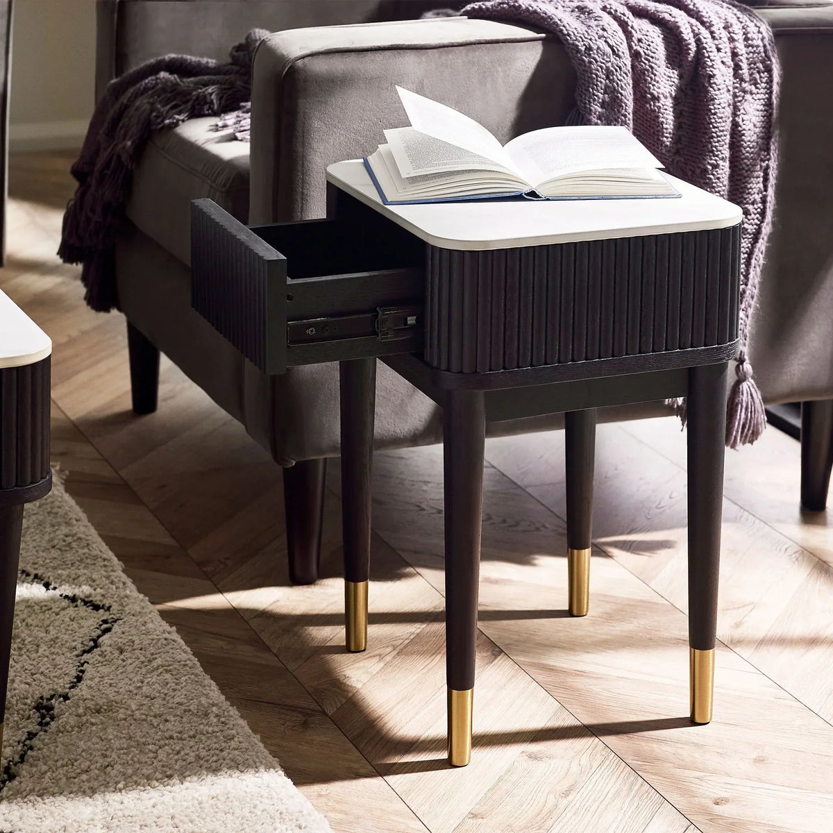 Black fluted lamp table with white stone top and open drawer beside grey sofa in living room