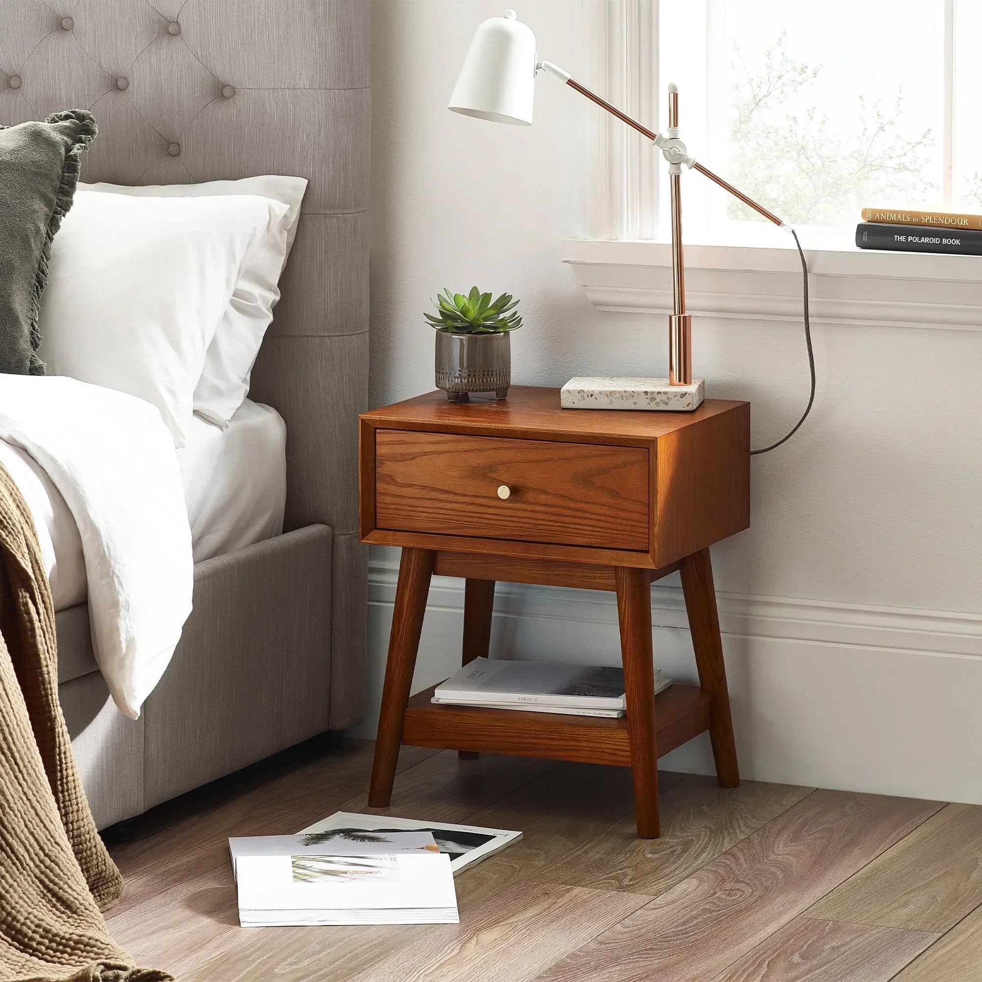 Walnut one drawer side table beside bed with lamp, plant, and books on shelf and floor