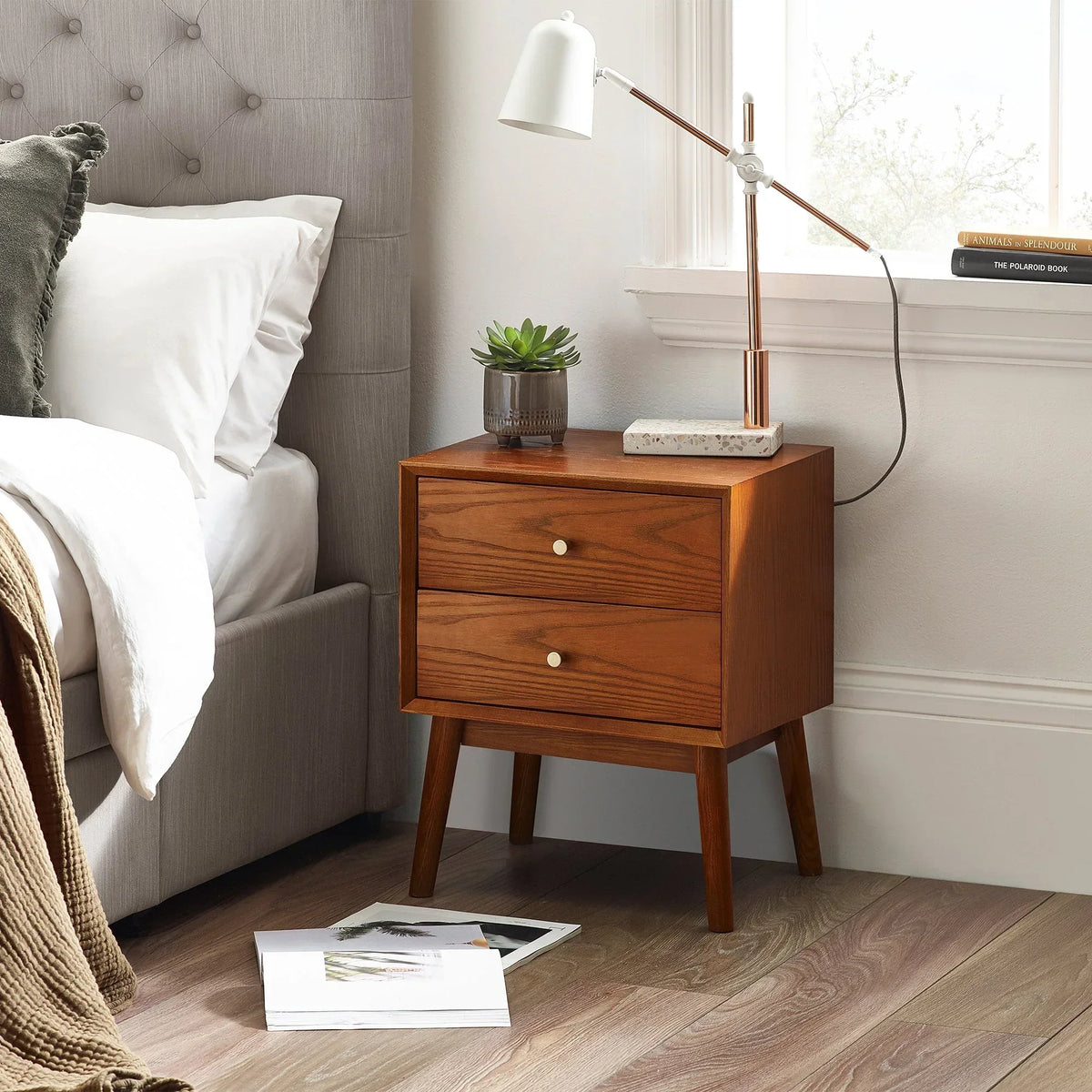 Mid-century oak bedside table with two drawers, lamp, and plant beside upholstered bed