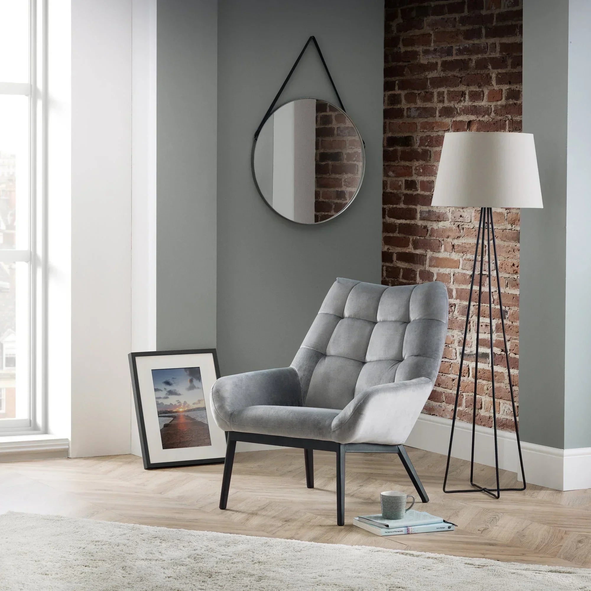 Grey velvet accent chair with black legs in modern living room with brick wall and floor lamp