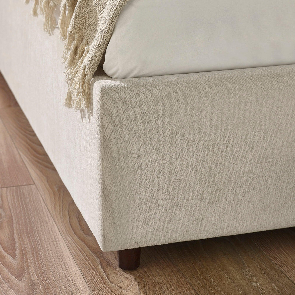 Beige upholstered bed base with wooden legs on oak flooring, close-up corner view