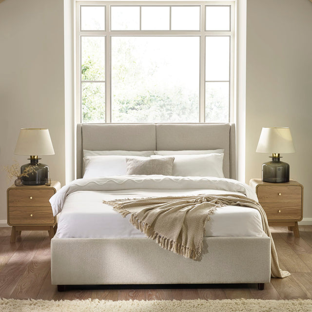 Beige upholstered bed with high padded headboard, white bedding, two wooden bedside tables with lamps