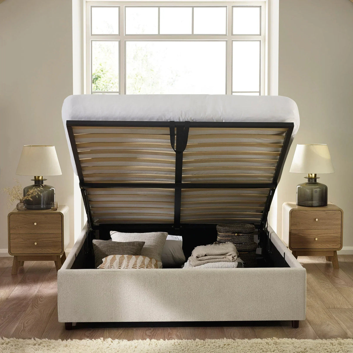 Beige upholstered ottoman storage bed with open lift-up base, pillows and blankets inside