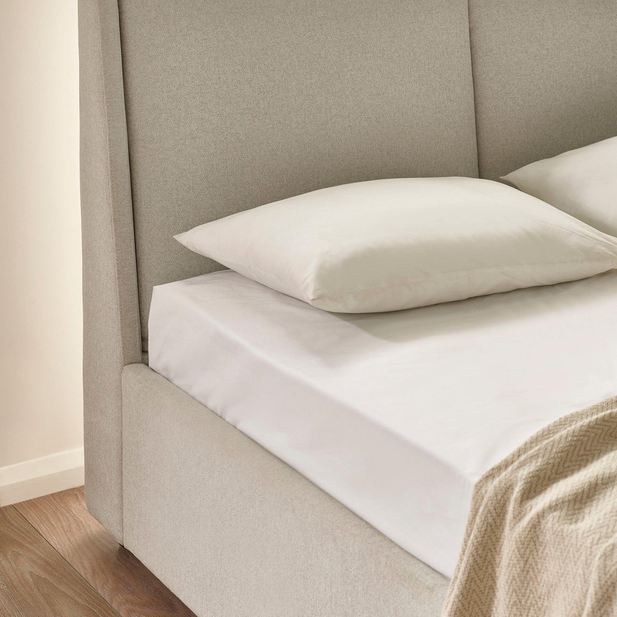 Beige upholstered bed frame with padded headboard, white bedding and pillow on wood floor