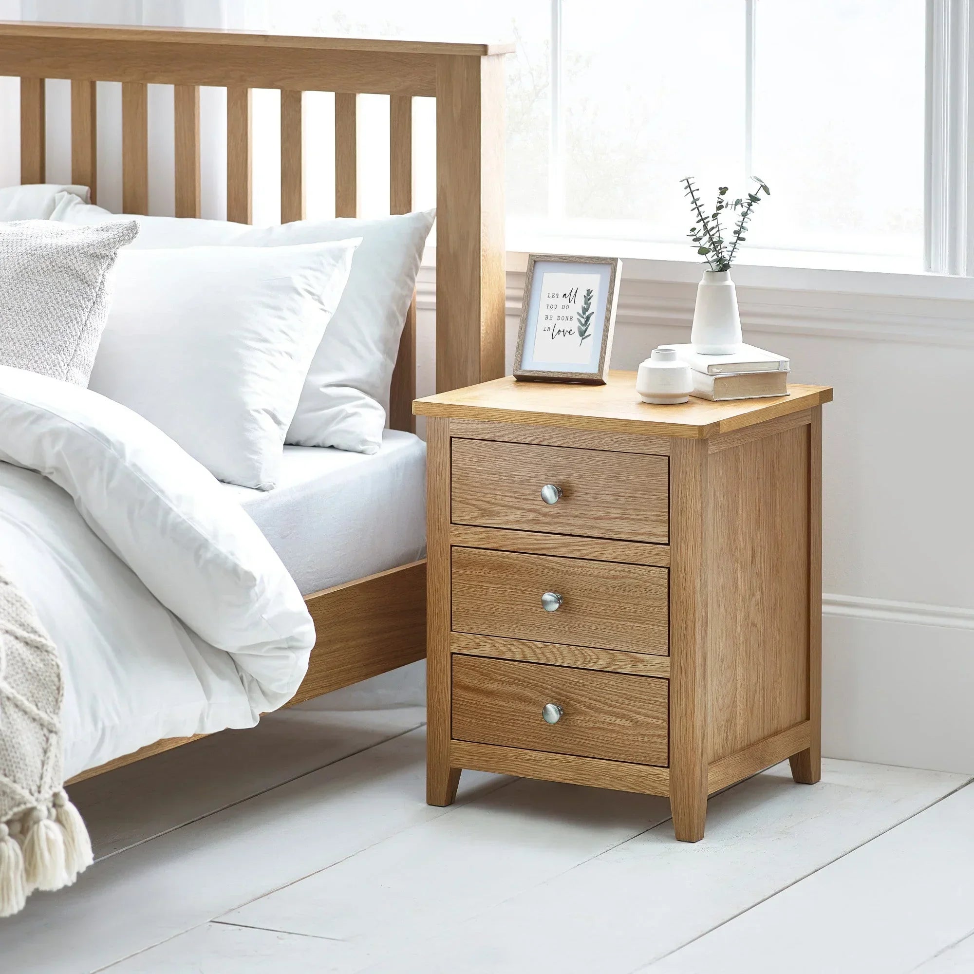 Oak three drawer bedside cabinet with metal handles beside bed in bright bedroom