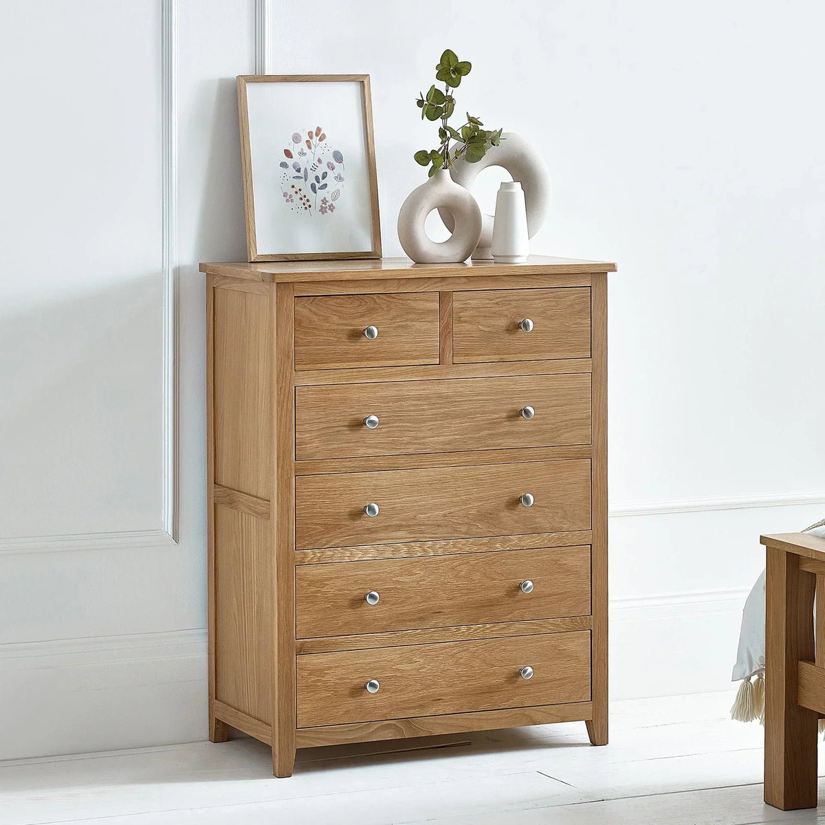 Oak chest of drawers with metal handles in bright bedroom, decorated with vases and artwork