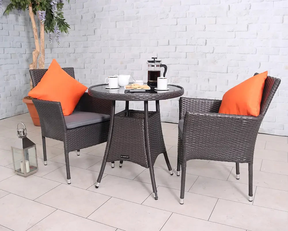 Brown rattan patio bistro set with glass top table, two chairs, orange cushions, coffee setup