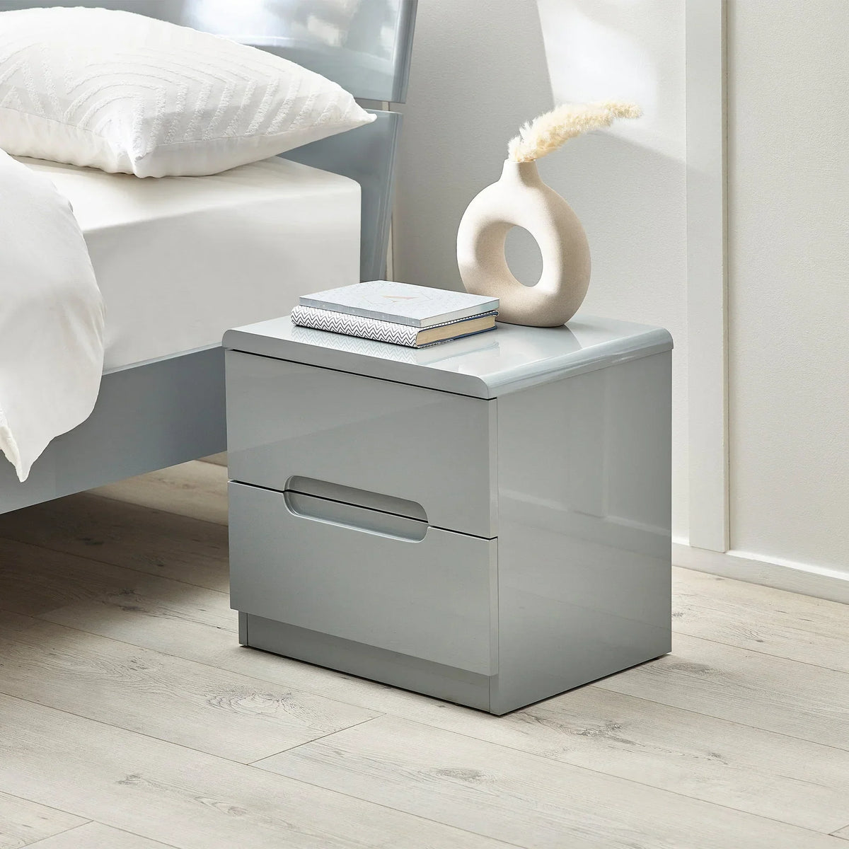 Grey two drawer bedside table beside bed with books and ceramic vase on wood floor
