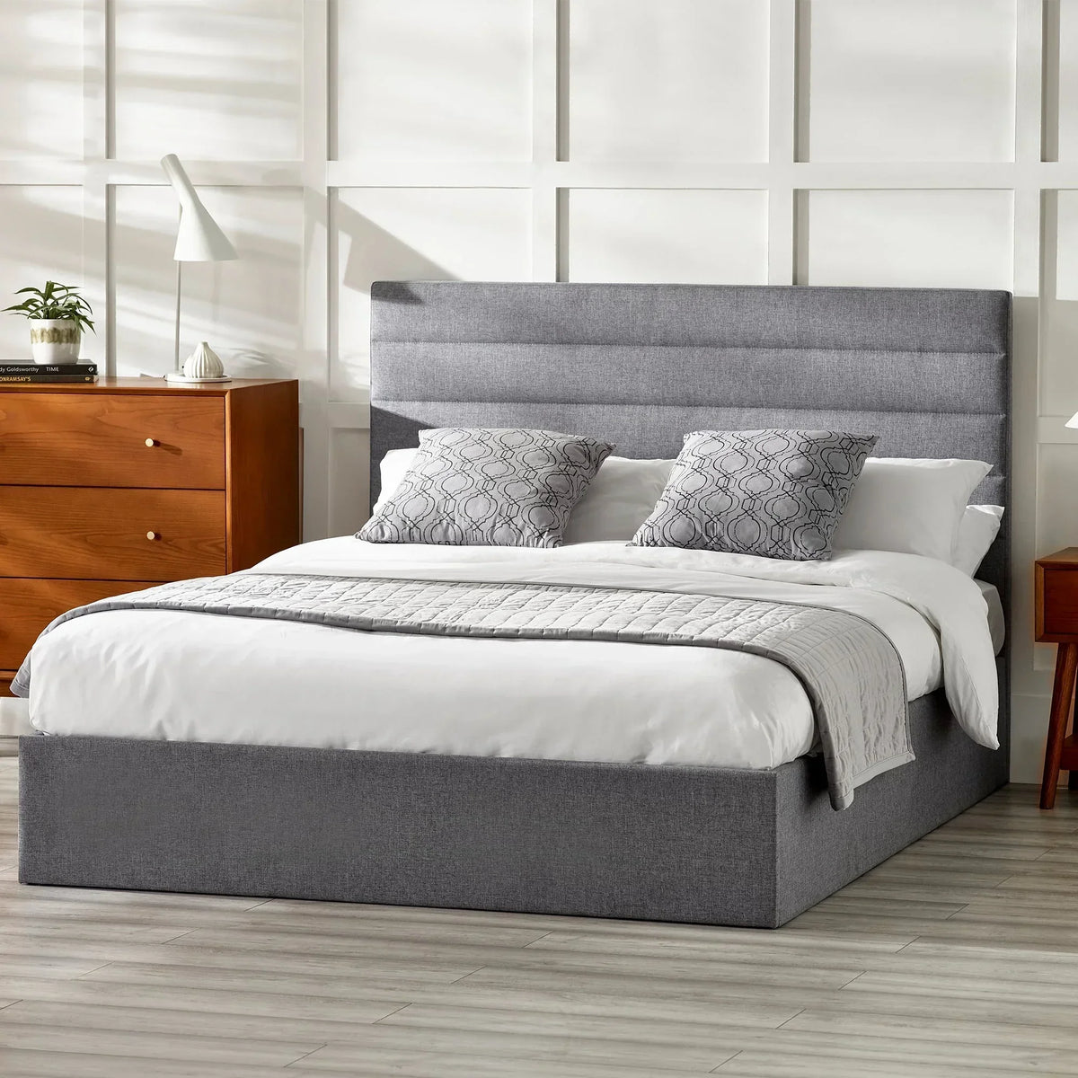 Grey lift-up storage bed with padded headboard, white bedding, and wooden drawers in bedroom