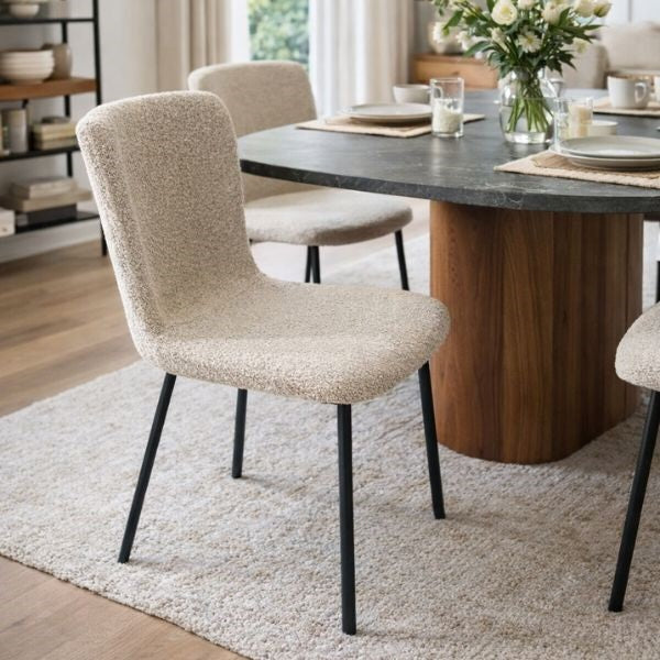 A modern dining room scene featuring a textured beige upholstered dining chair with black metal legs, set next to a round dark stone-top dining table with a wooden pedestal base.