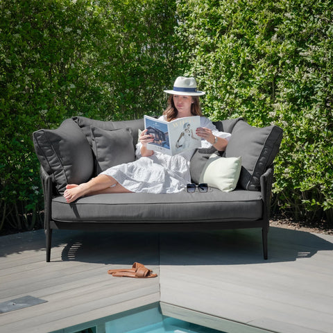 Dark grey outdoor sofa with plush cushions on a patio by pool, person reading magazine