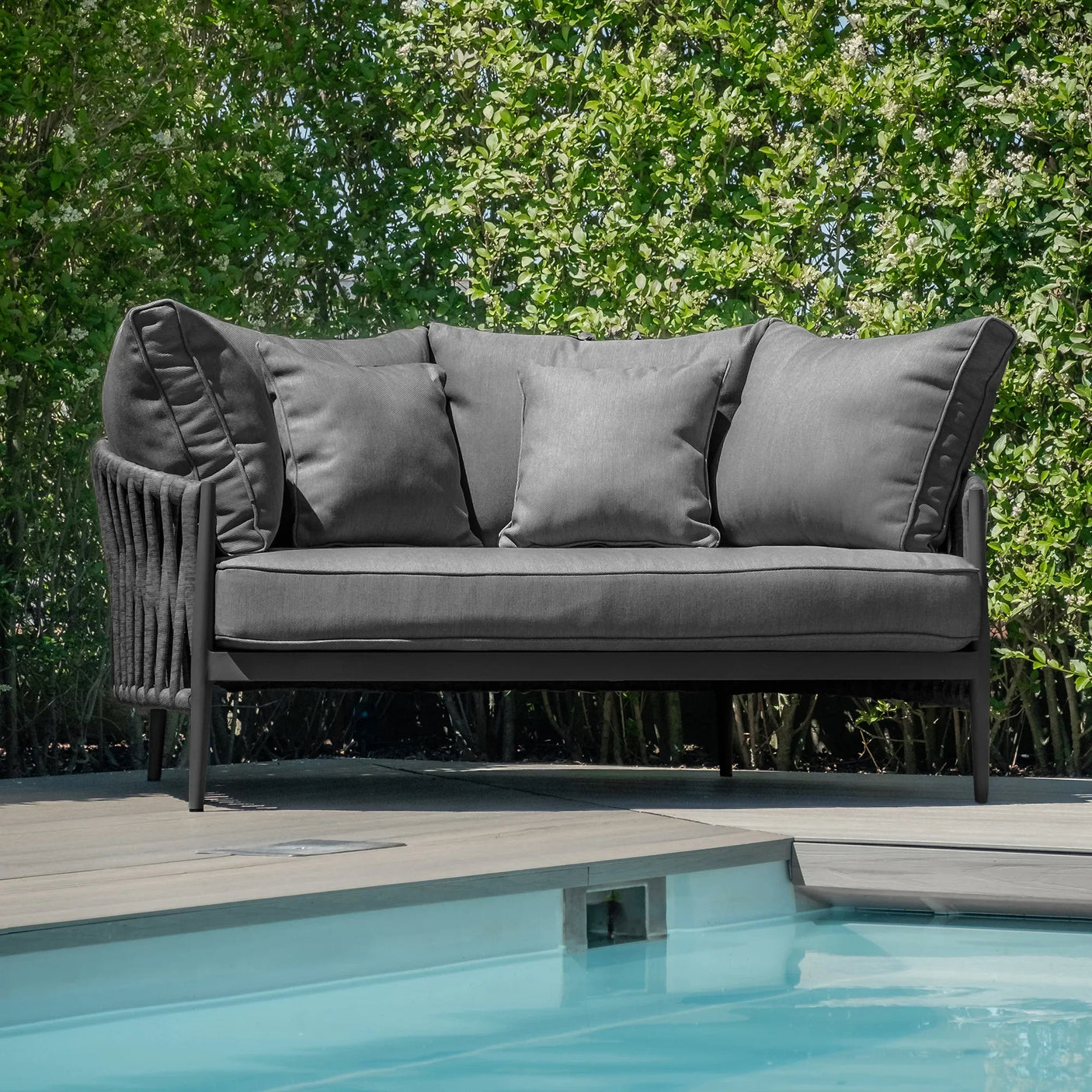 Grey outdoor sofa with cushions on patio beside swimming pool, surrounded by green hedges