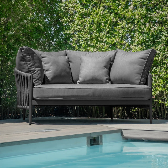 Grey outdoor sofa with cushions on patio beside swimming pool, surrounded by green hedges