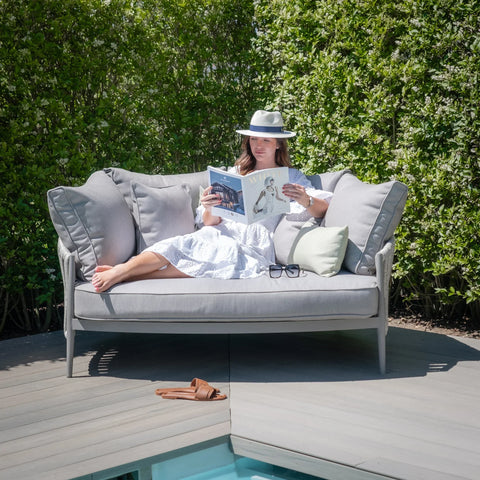 Grey outdoor sofa with plush cushions on deck by pool, woman reading magazine, green hedge backdrop