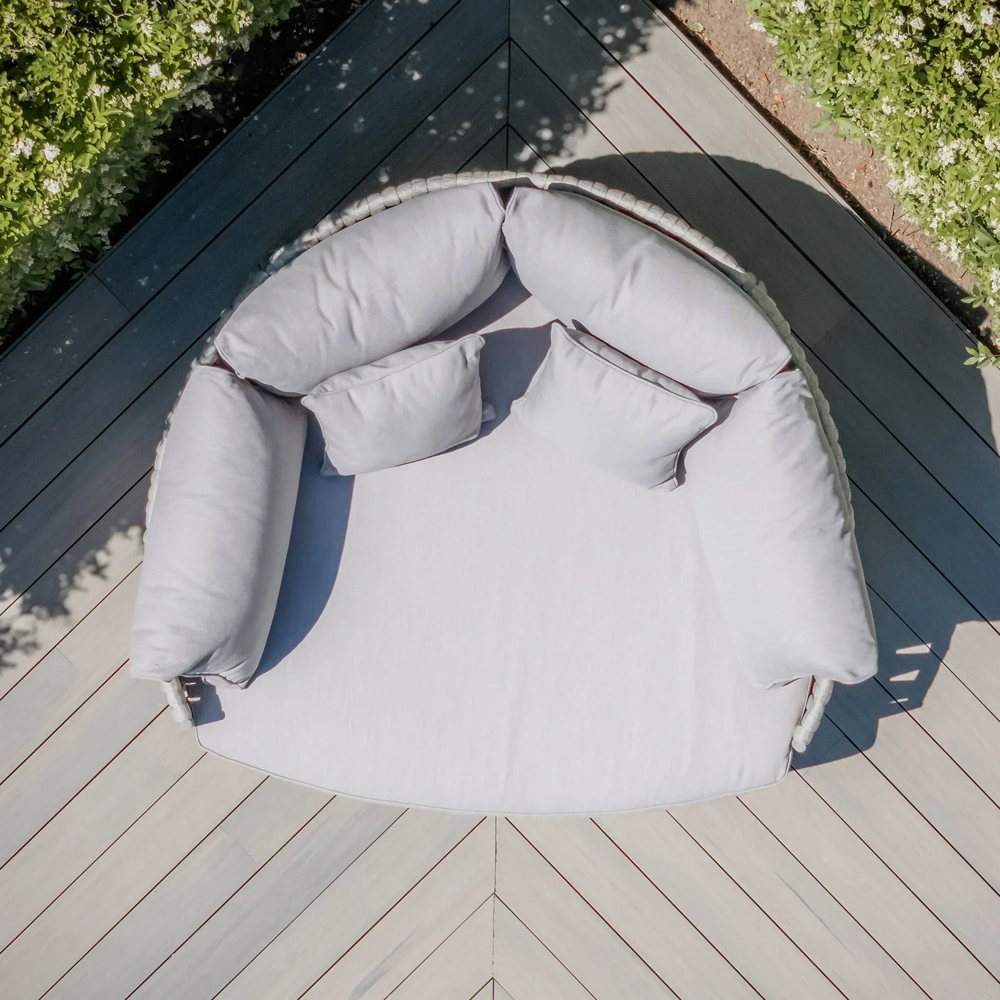 Round outdoor daybed with light grey cushions on wooden decking, aerial view
