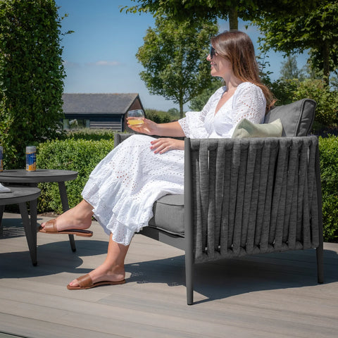 Grey outdoor lounge chair with rope detailing and cushions on patio, woman relaxing in sunlight