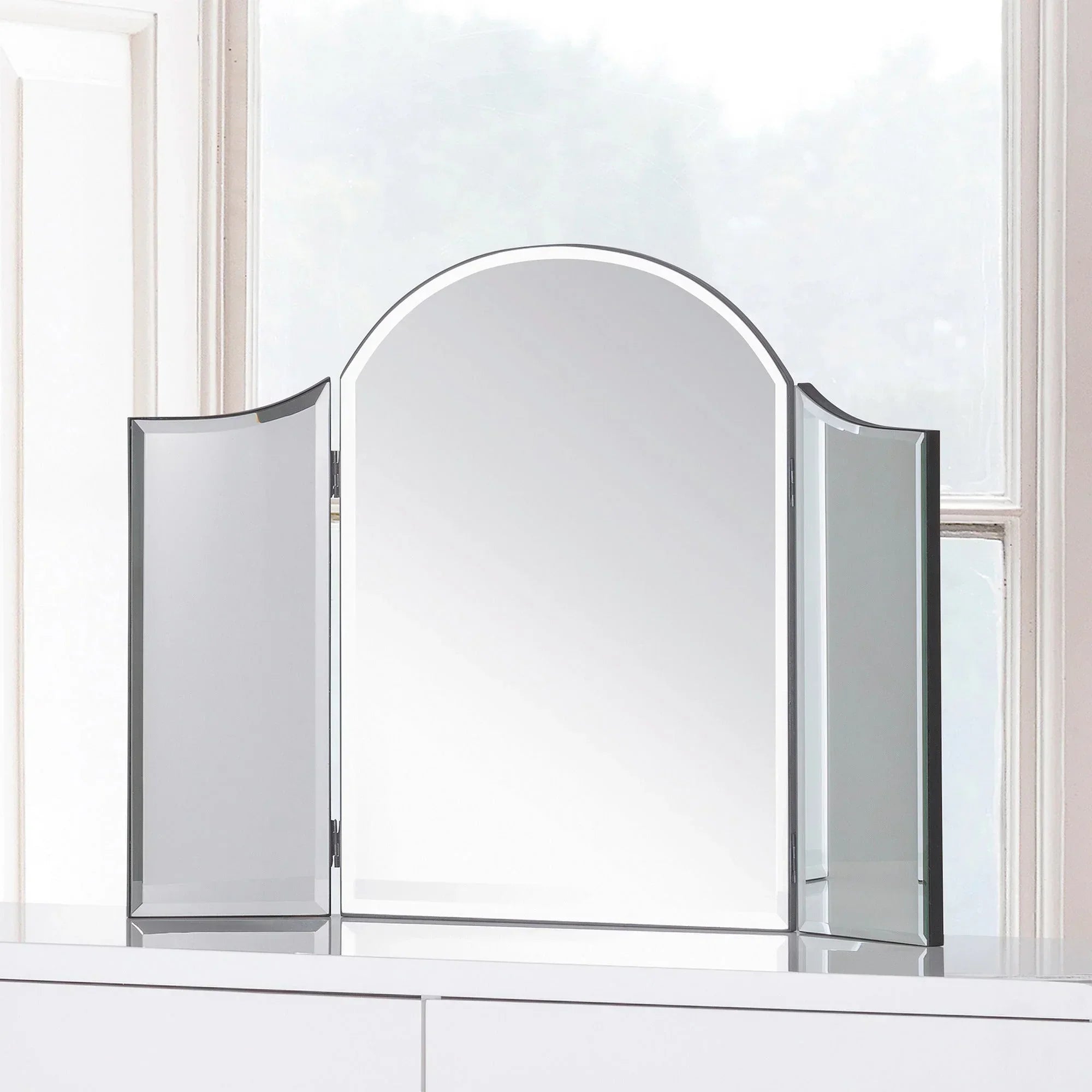 Curved dressing table mirror with three glass panels on a white vanity in front of a window