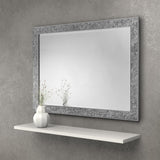 Rectangular wall mirror with decorative silver textured frame above a white floating shelf