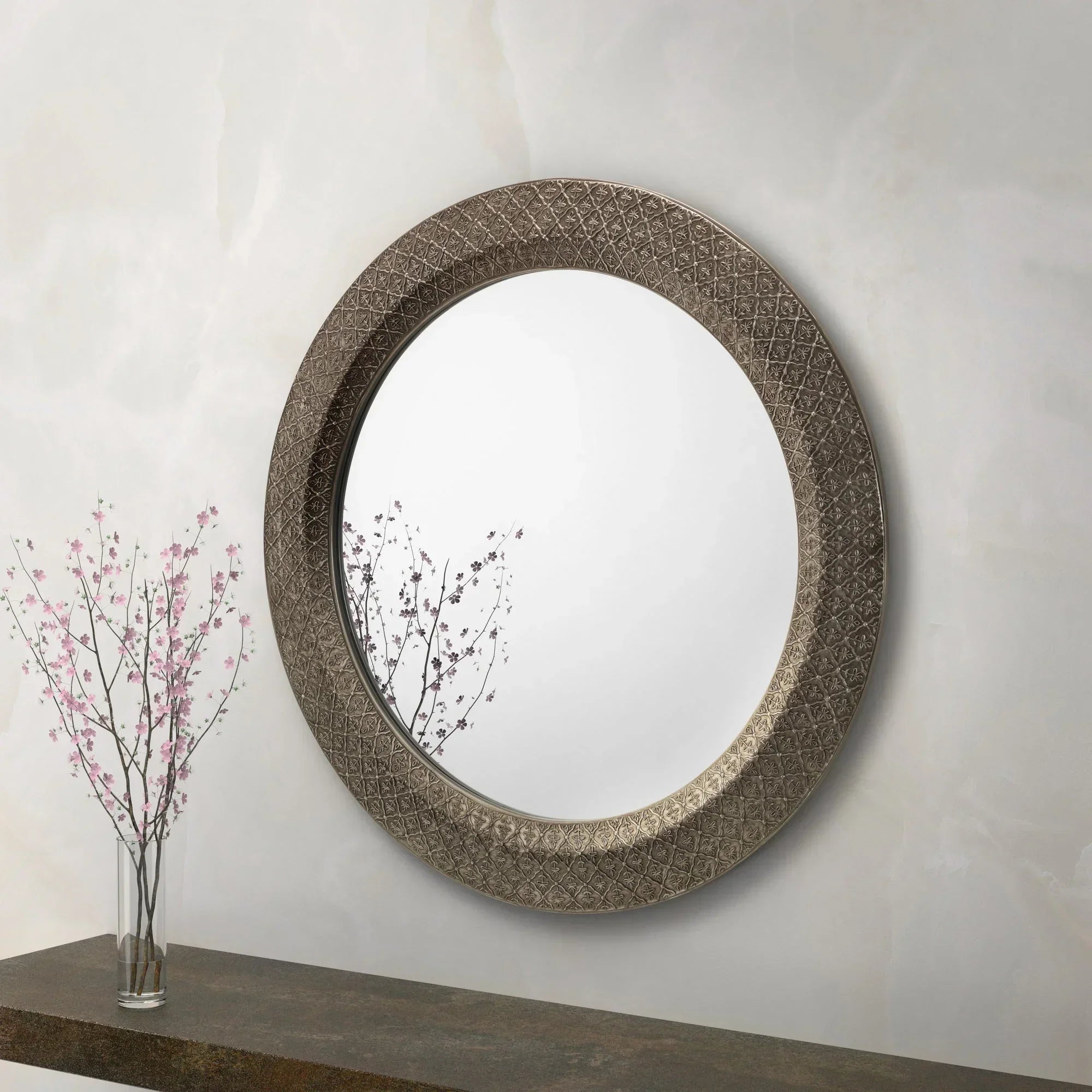 Large round mirror with ornate metal frame above console table and glass vase with pink flowers