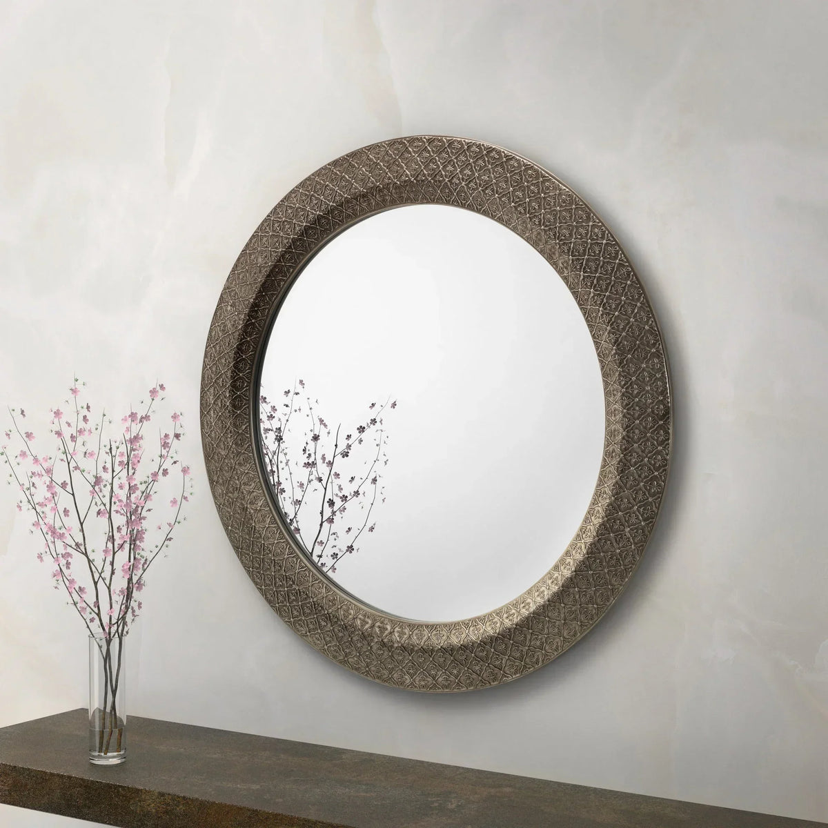 Large round mirror with ornate metal frame above console table and glass vase with pink flowers
