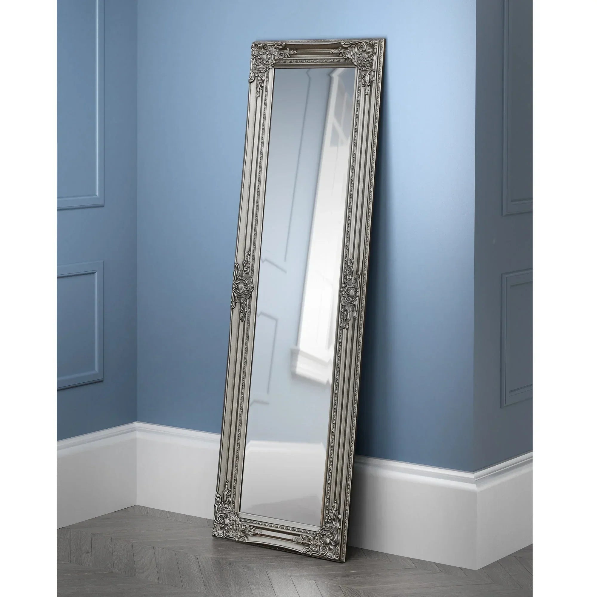 Full length pewter dress mirror with ornate frame leaning against blue paneled wall