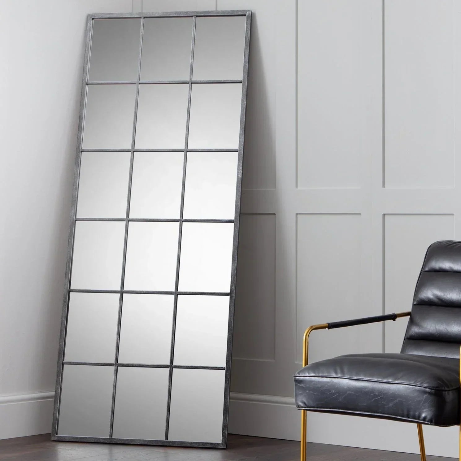 Tall windowpane mirror with black metal frame leaning against panelled wall, armchair beside