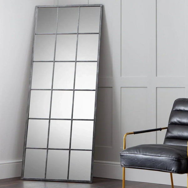 Tall windowpane mirror with black metal frame leaning against panelled wall, armchair beside
