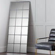 Tall windowpane mirror with black metal frame leaning against panelled wall, armchair beside