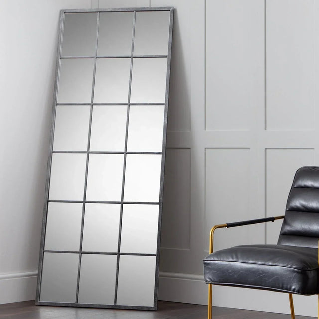 Tall windowpane mirror with black metal frame leaning against panelled wall, armchair beside