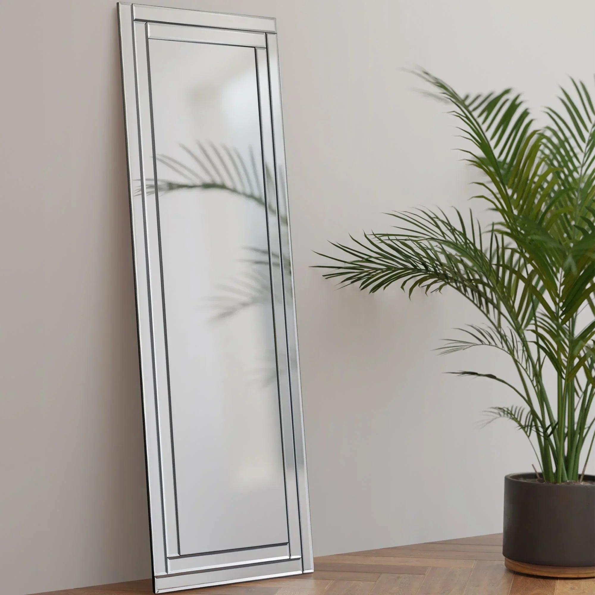 Frameless dress mirror with beveled edge leaning on wooden table beside potted green plant
