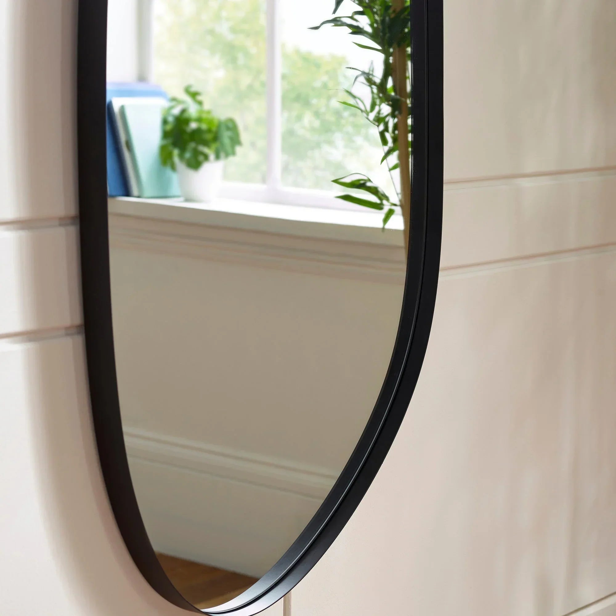 Oval wall mirror with black frame reflecting window and indoor plant