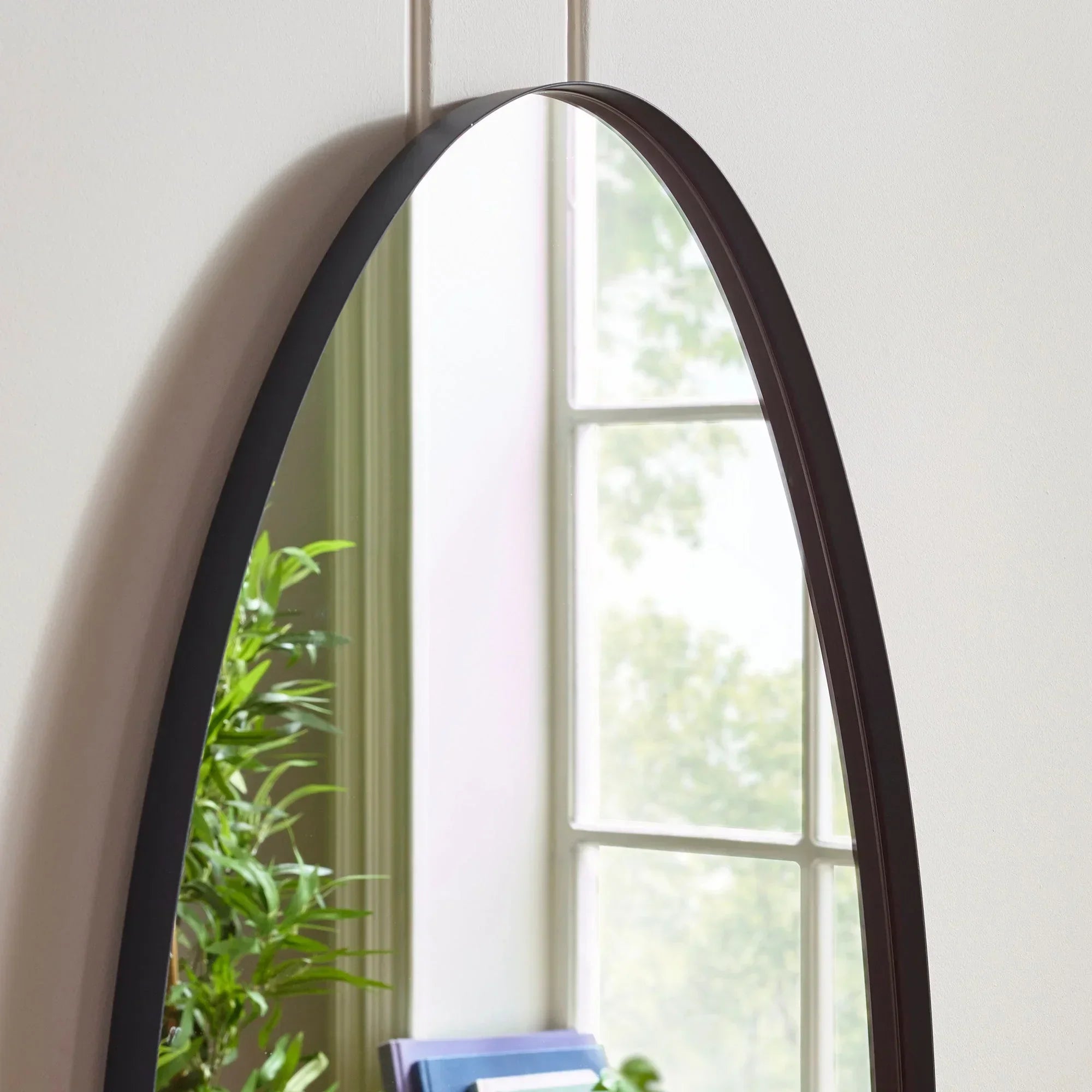 Arched wall mirror with slim black metal frame reflecting window and houseplant