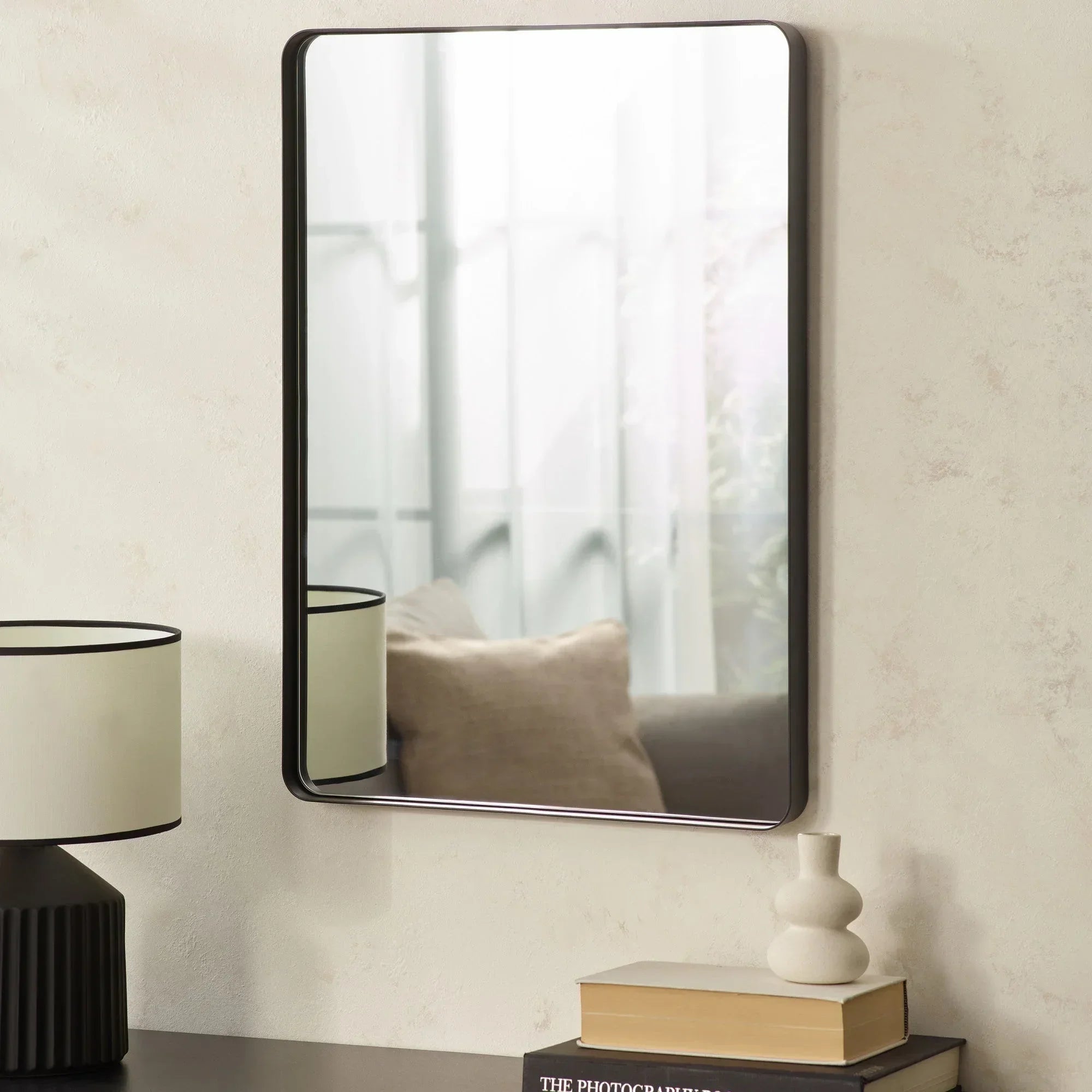 Rectangular wall mirror with black frame above side table, lamp, books and decorative vase