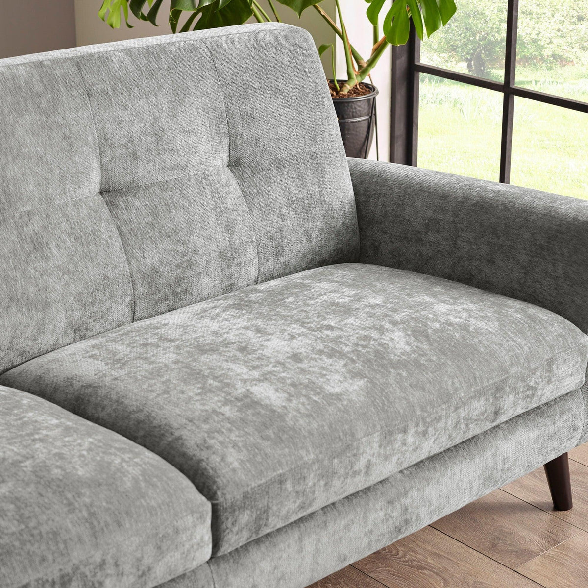 Grey fabric sofa with wide armrests and tufted backrest near window and plant