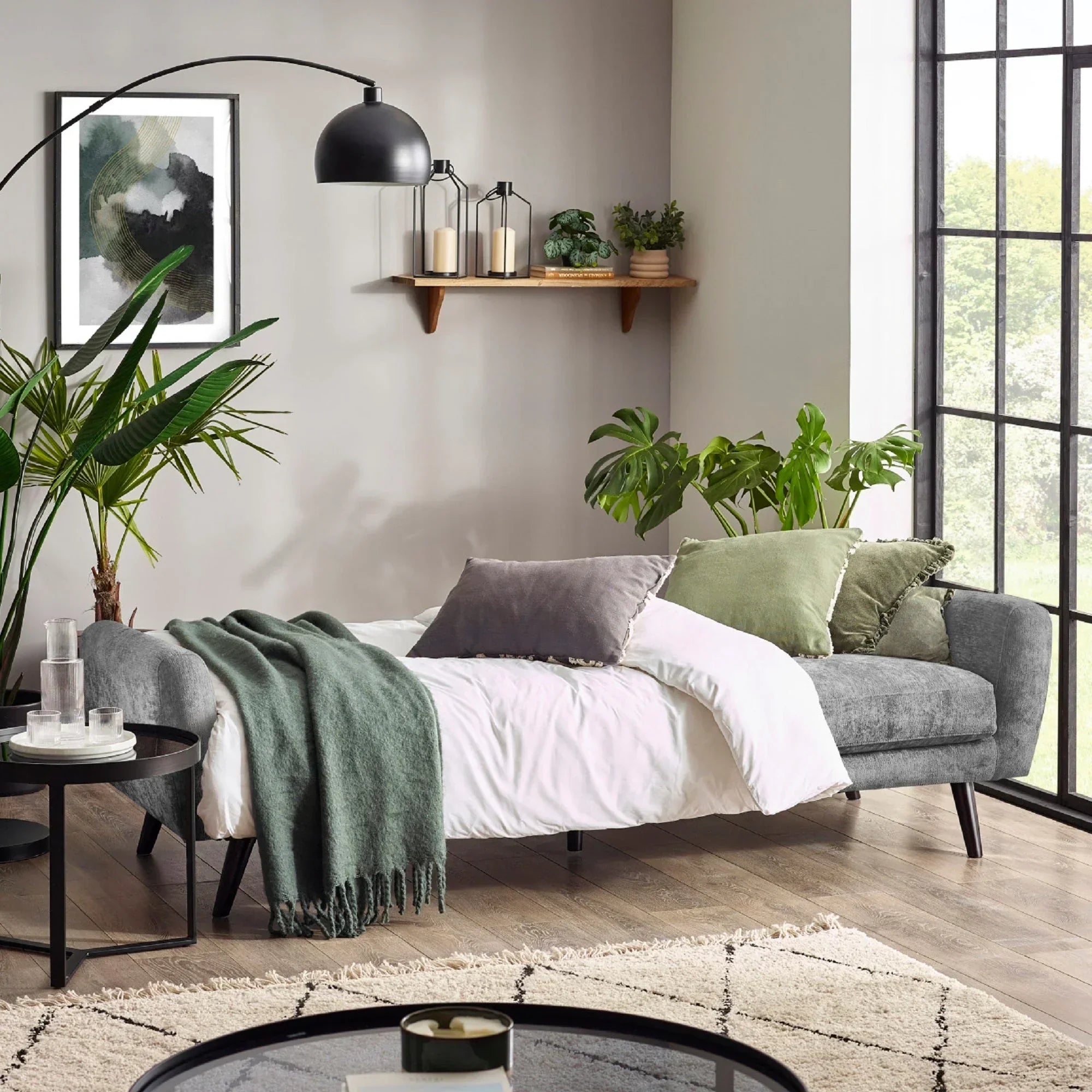 Grey fabric sofa bed with green and grey cushions in modern living room with plants and blanket